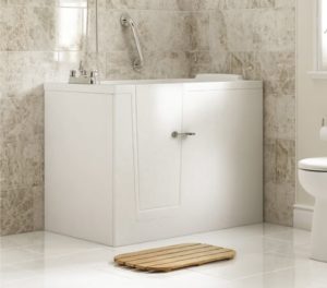 A modern walk-in bathtub with a closed door, a safety grab bar, and a faucet. A wooden bath mat is placed on the white tiled floor in front of the tub, and a toilet is partially visible on the right.