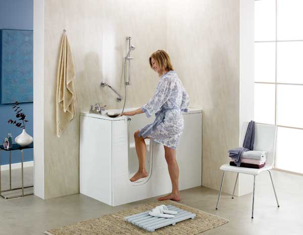 A woman in a robe steps into a walk-in bathtub in a modern, well-lit bathroom with a towel rack, chair with folded towels, and minimalist decor.