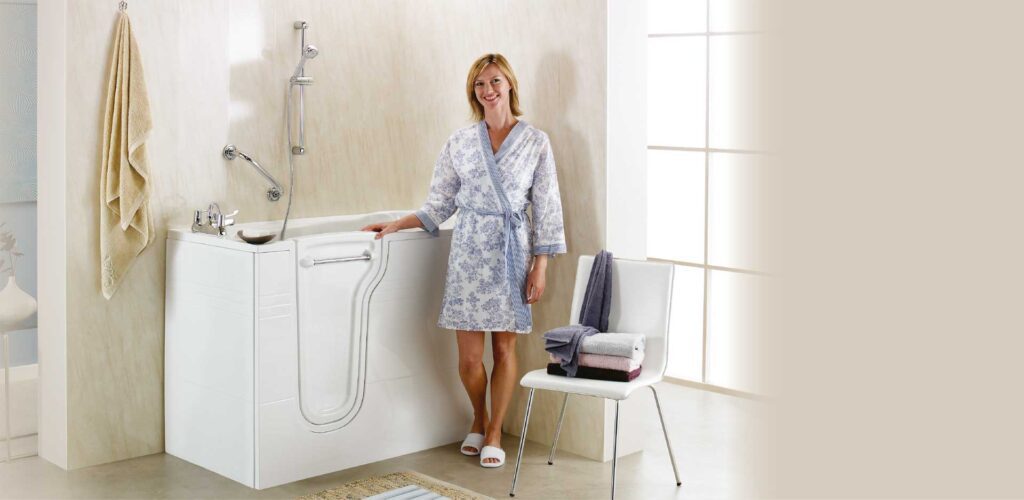 A smiling woman in a robe stands beside a modern walk-in bathtub in a bright bathroom. A chair with neatly folded towels is nearby, and a towel hangs on the wall.