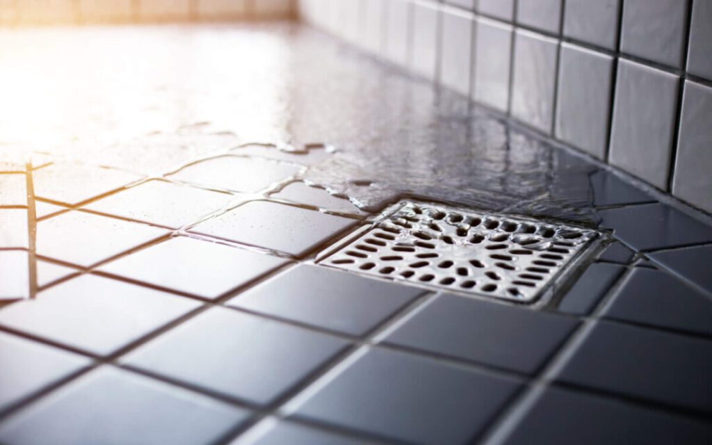 A close-up of water pooling on a tiled floor near a metal drain, with some water spilling over the tiles and reflecting light.