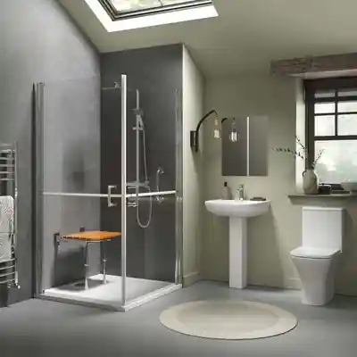 Modern accessible bathroom with walk-in shower featuring clear glass doors, a fold-down wooden seat, and grab rails for safety. The space includes a white pedestal sink, a toilet, neutral grey walls, a round beige rug, and a window for natural light.