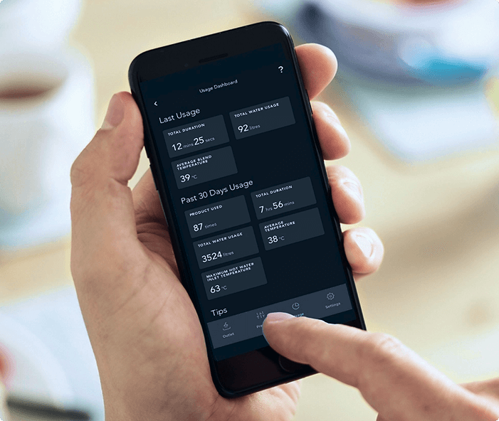 Alt text: A hand holding a smartphone showing a usage dashboard app screen. The app displays data including last usage duration (12 mins 25 secs), total water usage (92 litres), average end temperature (39°C), past 30 days usage with total duration (7 hrs 56 mins), product used (87 times), total water usage (3524 litres), average temperature (38°C), and maximum hot water inlet temperature (63°C). A finger is about to tap on an icon at the bottom of the screen labeled “Tips.” The background is blurred with a person and a table visible.