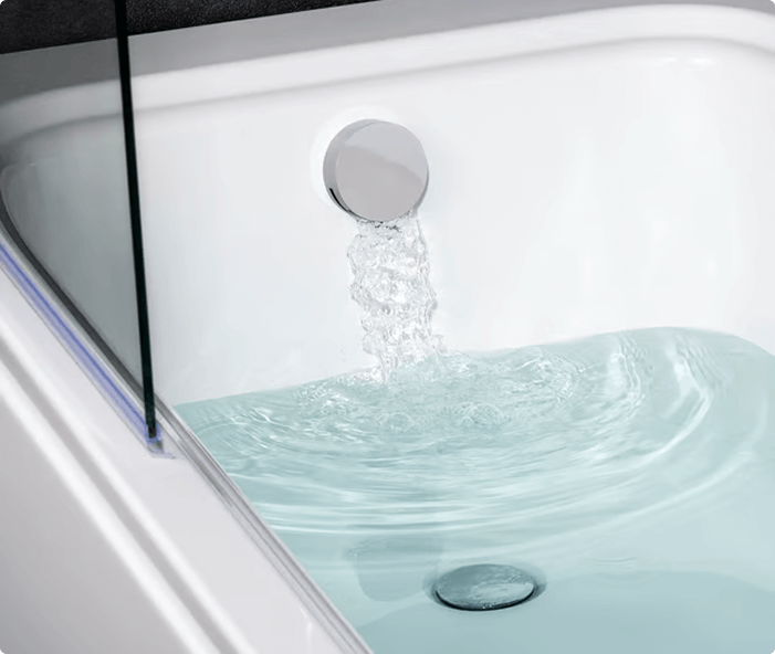 Close-up of water flowing from a circular chrome bath filler into a white bathtub filled with clear water, with a glass shower screen visible on the left side.