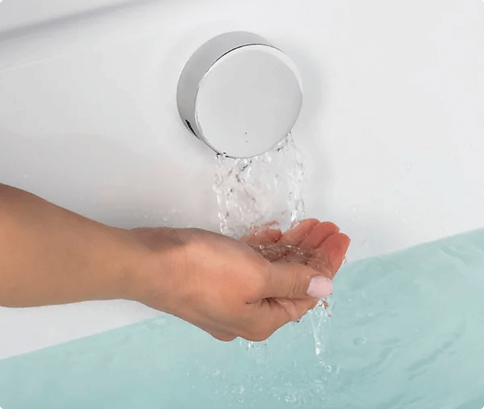 Person’s hand cupped under water flowing from a round chrome bath filler into a white bathtub filled with clear water.