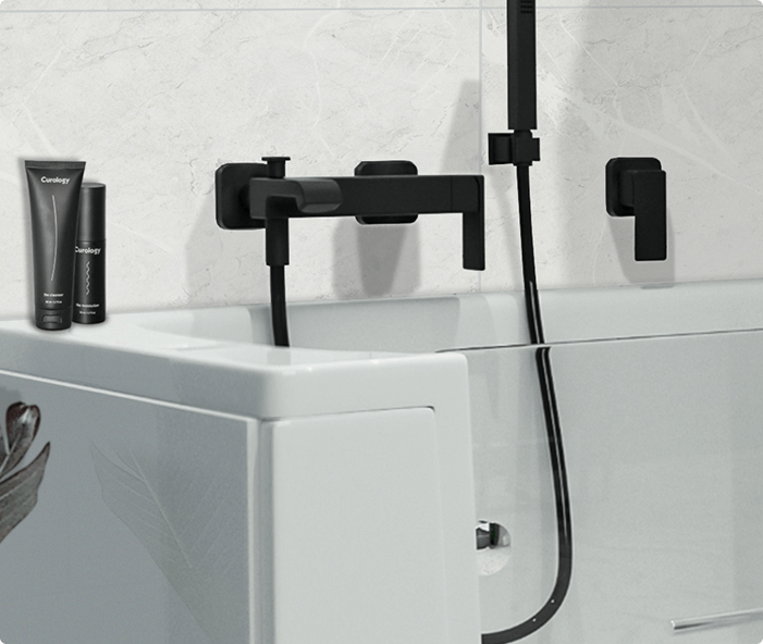 This is the Hampton Elite bath, featuring sleek black fixtures including a bath filler, shower handset, and control lever mounted on the wall above the bath. The bath has a clear walk-in door with a white frame, blending modern design with user-friendly accessibility. The image shows a close-up of the bath corner with some toiletries on the edge, highlighting both functionality and style.