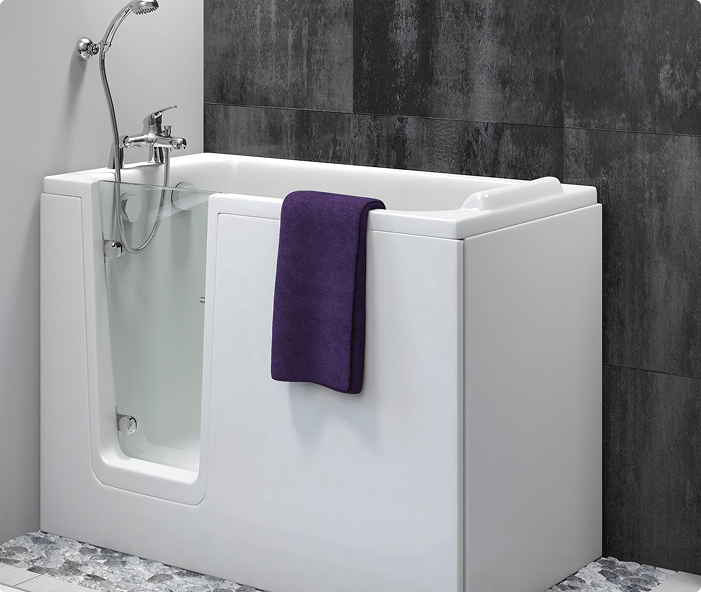 This is another view of the Hampton Elite bath. It features a white walk-in bath with a clear glass door and chrome hinges. There is a built-in seat inside the bath, a chrome bath filler with a shower handset, and a purple towel draped over the side for contrast. The bath is set against a dark tiled wall, creating a modern and stylish bathroom look. The pebble-effect flooring adds a natural texture to the space.