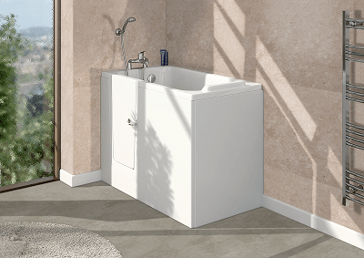Contemporary accessible bathroom showcasing a sleek white walk-in bath with a side-opening door and built-in seat, positioned beside a large window. The space features beige stone-effect tiled walls, grey flooring, a silver heated towel rail, and a round light grey rug.