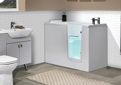 Modern accessible bathroom featuring the Hampton walk-in bath with a side-opening door and built-in seat, finished in white. The bathroom also includes a grey vanity unit with basin, a white toilet, white subway tile walls, and a beige patterned rug on the floor.