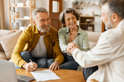 Older couple seated at a table with documents, consulting with a professional. Man is holding a pen, suggesting review or signing.