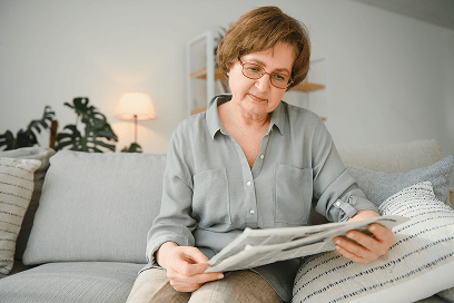 Older woman sitting on a couch reading a newspaper in a cozy, well-lit living room.