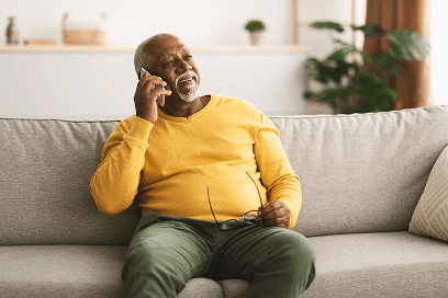 Older man smiling while talking on phone, sitting on a sofa at home holding eyeglasses in his other hand.