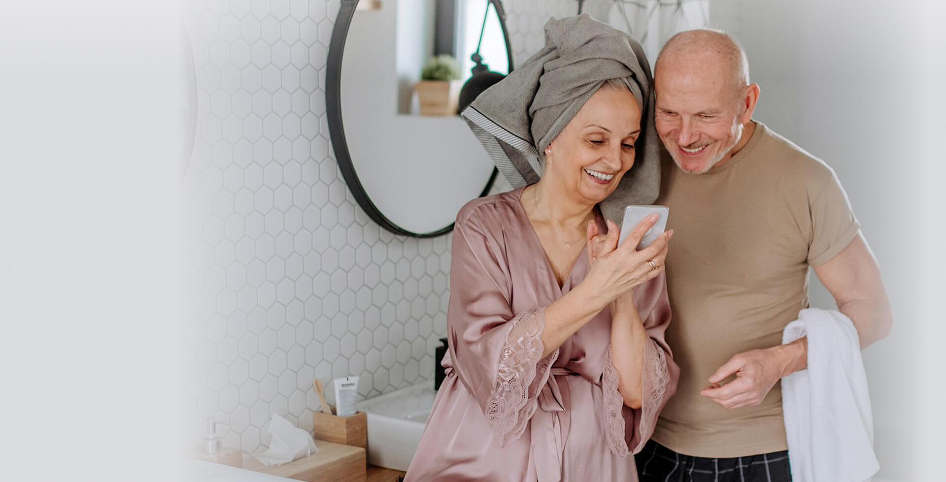Smiling older couple in a bright bathroom, looking at a smartphone together after a shower, with a towel and robe