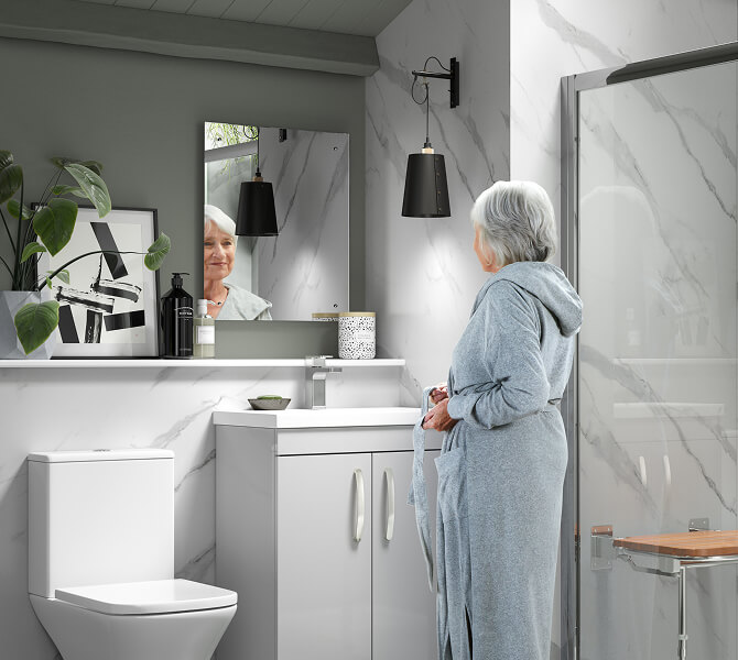 Older woman in a gray bathrobe looking into a bathroom mirror above a white vanity unit, surrounded by white marble walls, a toilet, and a glass shower enclosure with a wooden seat.