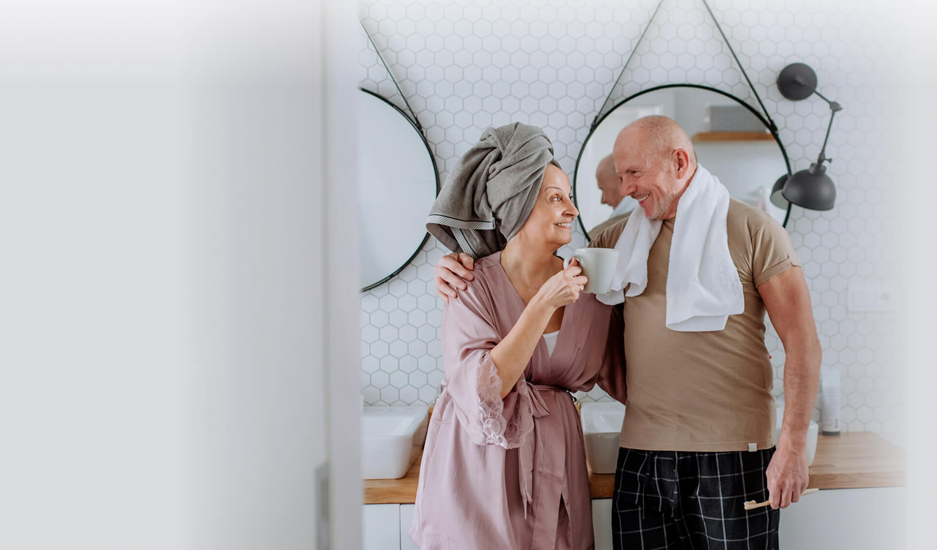 Older couple smiling at each other in a bathroom, wearing loungewear with towels over their shoulders, sharing a relaxed moment by the sink