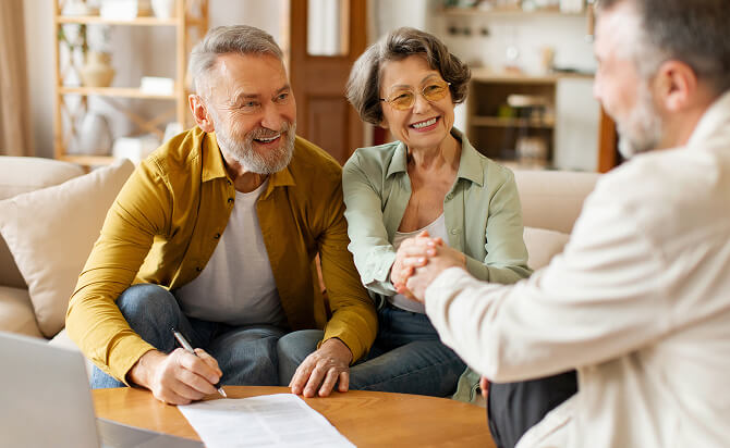Older couple smiling warmly while signing documents at home, with a man in a light-colored suit shaking hands across a wooden table in a bright, cozy living room.