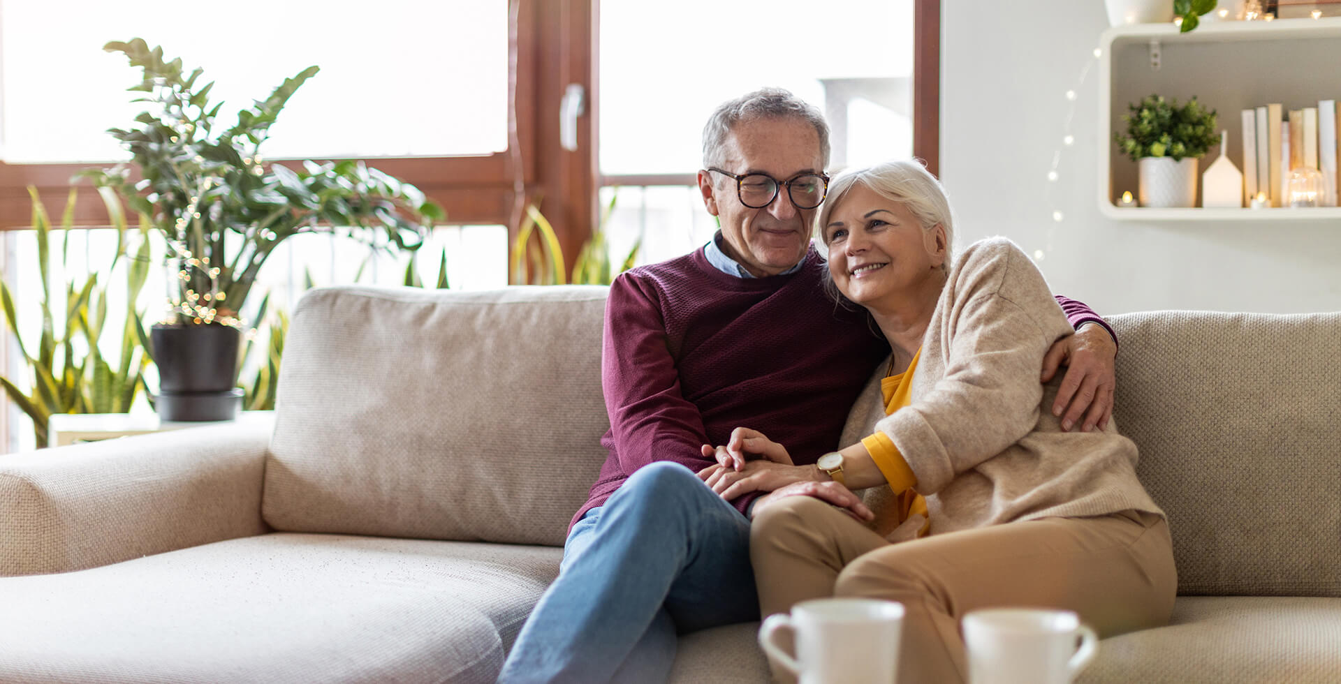 Older couple sitting close together on a beige sofa, smiling and holding hands in a cosy living room with houseplants and soft lighting
