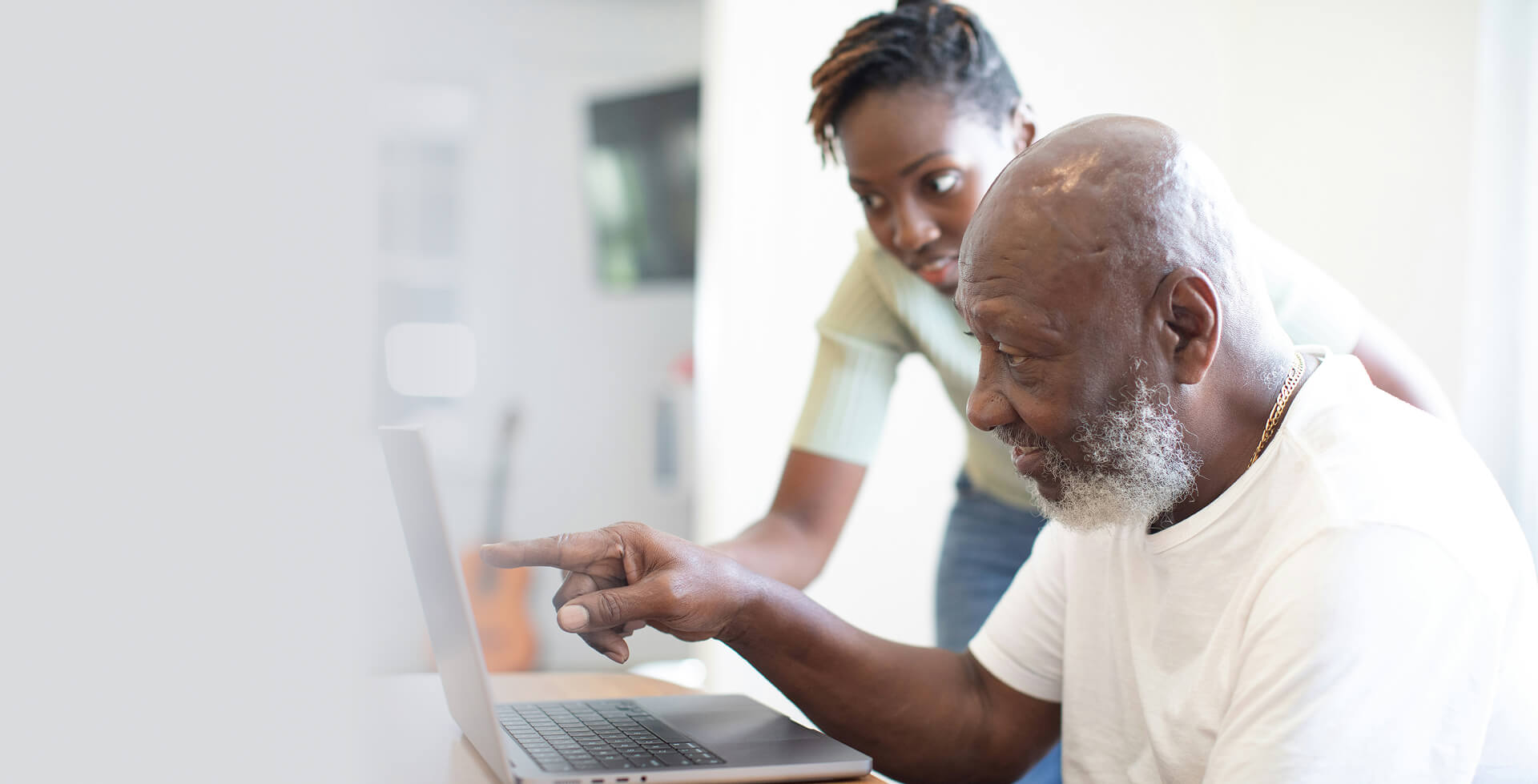 Older man using a laptop with support from a younger woman in a bright, modern home setting