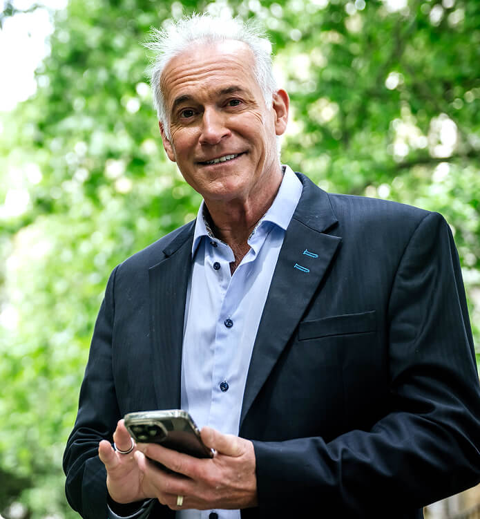 Dr. Hilary Jones, a distinguished medical doctor and TV presenter, smiling and holding a smartphone, standing outdoors with green trees in the background, dressed in a dark blazer and light blue shirt.
