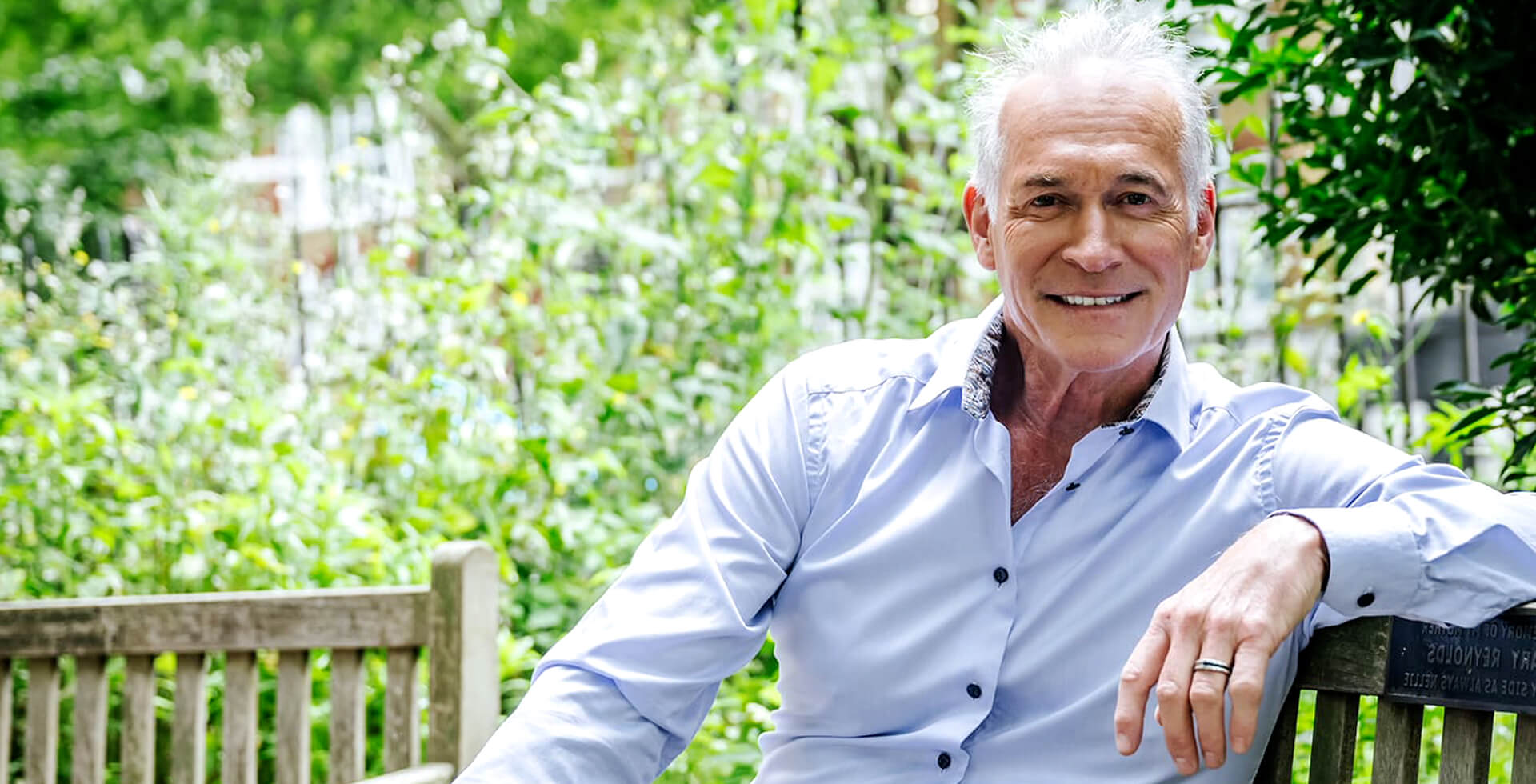 Dr Hilary Jones MBE seated on a garden bench, smiling at the camera in a relaxed outdoor setting. Best known for his work as a television doctor and health commentator.