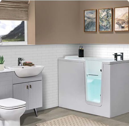 A bright bathroom featuring the Hampton Elite walk-in bath with a clear door and blue water inside, set against white subway tiles and a beige wall with three framed artworks. Nearby are a white toilet and a sink with cabinetry.