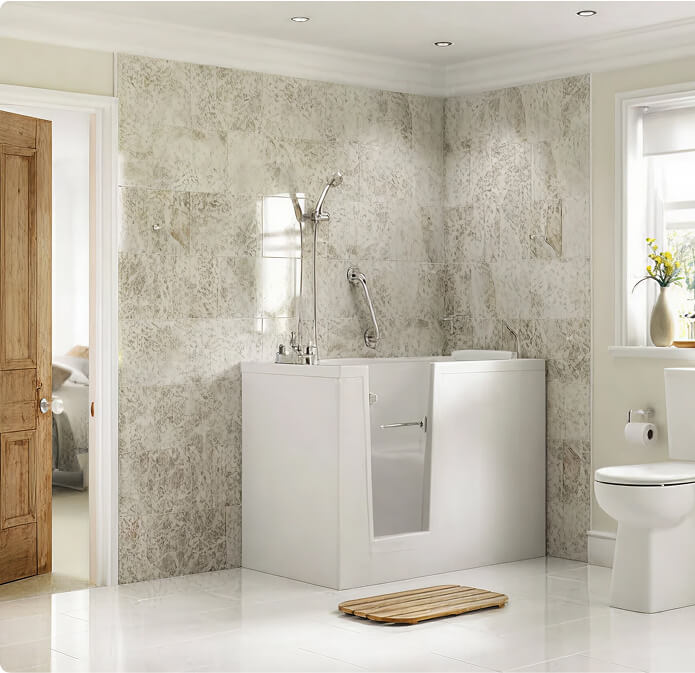 Walk-in bathtub with side access door, silver tiled walls, handheld shower, stainless steel grab bar, wooden bath mat, white floor tiles, white toilet, wooden door, and window with roller blind.