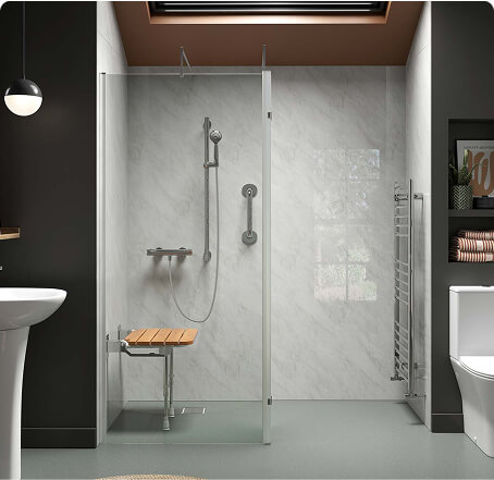 Accessible shower enclosure with glass walls, marble-effect walls, fold-down wooden seat, stainless steel grab bar, hand shower, pedestal sink, white toilet, grey floor, and a skylight ceiling.