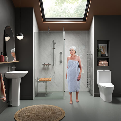 A modern accessible bathroom with dark walls and a skylight. A woman wrapped in a towel stands barefoot on a smooth grey floor near a walk-in shower with a glass door, a fold-down wooden seat, and silver grab bars. To the left is a white pedestal sink with a large arched mirror and pendant lights, while on the right is a white toilet and a chrome towel radiator. A round woven rug lies on the floor.
