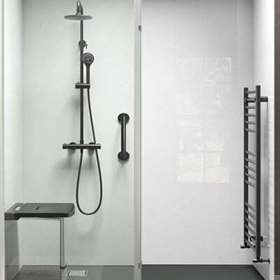 Shower area with matte black fittings, including an adjustable shower head, a built-in grab bar, and a foldable seat. Adjacent to the shower, there is a matching matte black heated towel rail on a white wall. The floor is dark grey with a minimalist design.