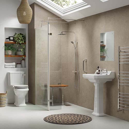 A spacious wetroom bathroom with beige walls and a large skylight. The room includes a walk-in shower with a glass enclosure and a fold-down wooden seat. There is a silver grab bar on the shower wall, a white pedestal sink with a mirror above, and a white toilet with a black striped seat cover. To the right, a tall chrome towel radiator is mounted on the wall. Built-in shelves with plants and towels add a touch of greenery, and a round patterned rug lies on the light floor.