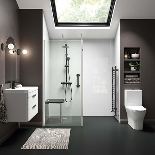 Modern accessible bathroom with dark walls and a large skylight. Features include a floating white vanity with two drawers, a wall-mounted mirror with two round lights, and a walk-in shower with clear glass doors, a fold-down black seat, a black showerhead, and grab bars. A white toilet sits beside a recessed shelving unit holding towels and decor. The floor is dark with a patterned rug in front of the shower.