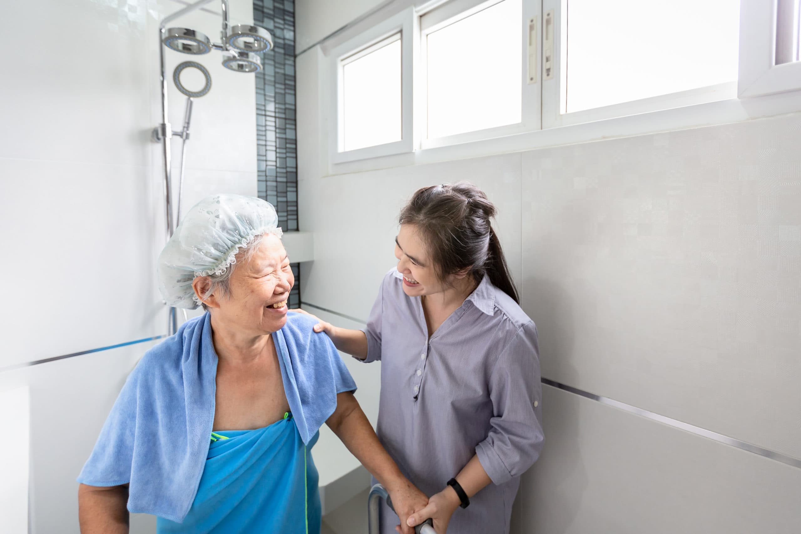 A smiling elderly woman wearing a shower cap and towel is helped by a younger woman in a bathroom.