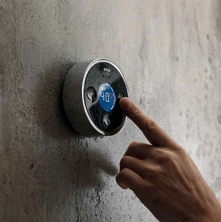 A hand adjusting a sleek, circular digital shower control set to 40°C, mounted on a textured grey wall with water droplets visible on the control.