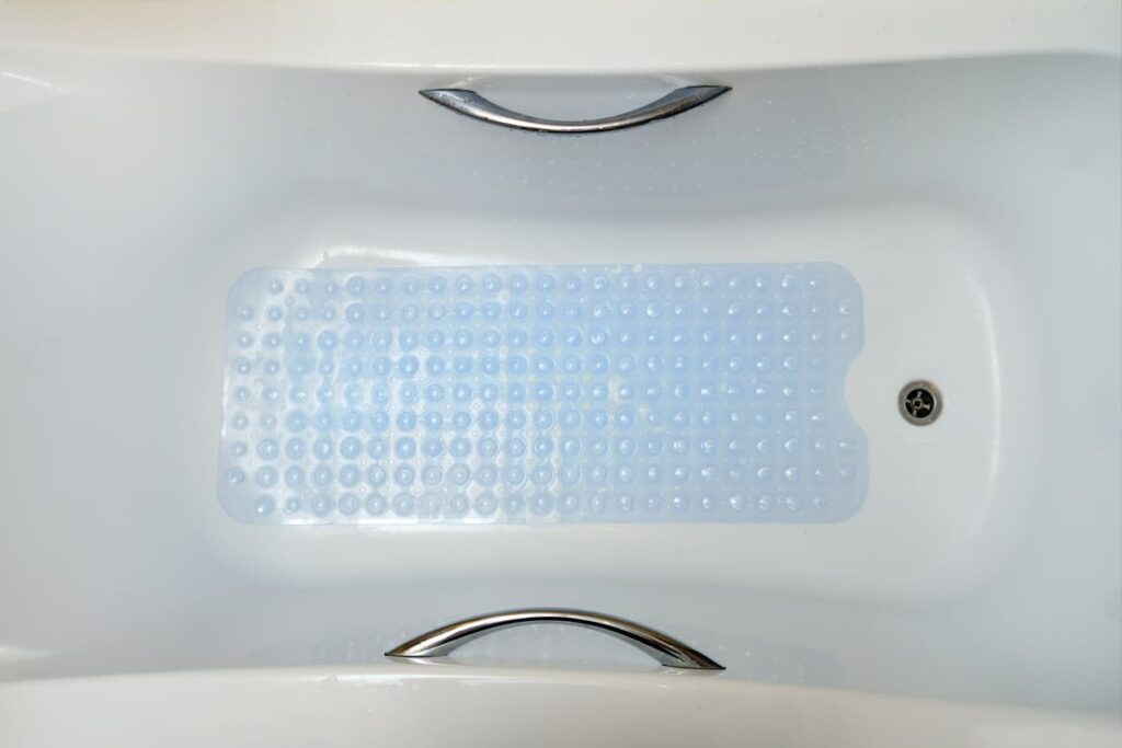 Top-down view of a white bathtub with a clear blue non-slip bath mat inside and two silver grab handles on the tub edges.